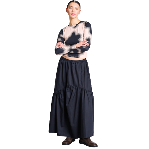 A woman stands with arms crossed, wearing a sheer, patterned long-sleeve top and a high-waisted, tiered black maxi skirt with brown boots. The background is white.