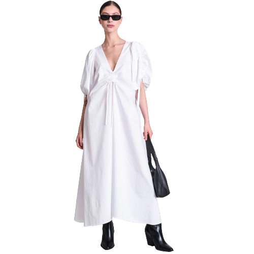 Woman in sunglasses wears a white, V-neck, puff-sleeve maxi dress with a front tie, black boots, and holds a black handbag, standing against a white background.